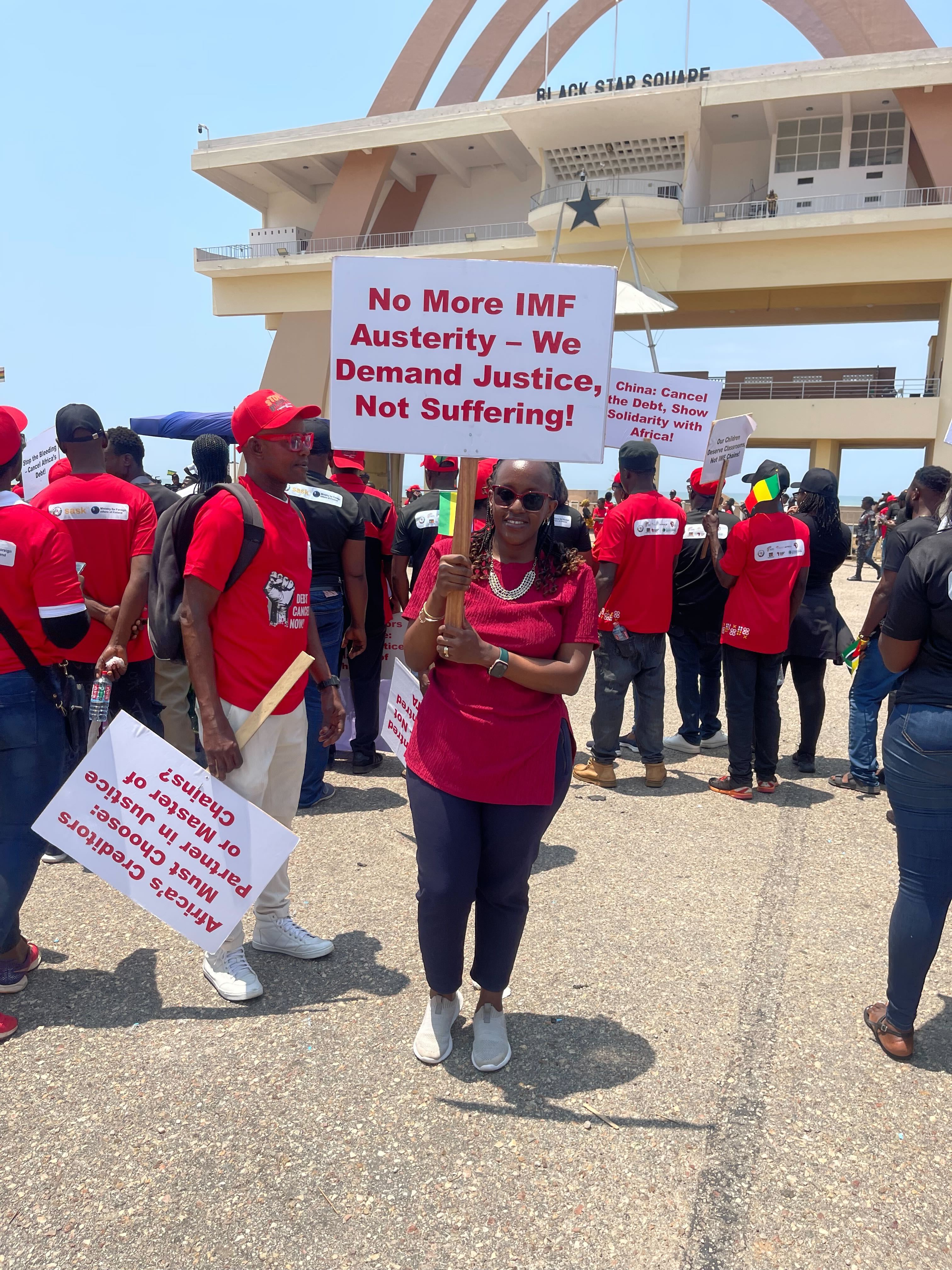 Peninnah Mbabazi stands at the front of a crowd during the Pan-Africa Debt Rally at Black Star Square in Accra, Ghana. She holds a sign that reads, “No More IMF Austerity – We Demand Justice, Not Suffering!” Other participants in red shirts hold protest signs in the background.